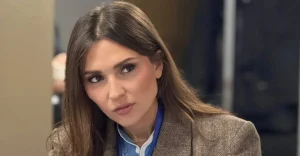 Жінка з довгим каштановим волоссям у твідовому піджаку та блакитній сорочці