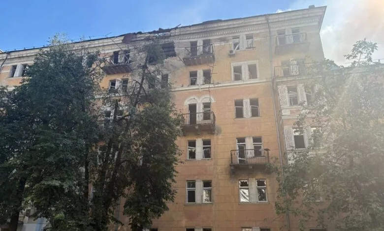 Пошкоджена жовта багатоповерхівка з вибитими вікнами та зруйнованим дахом серед дерев