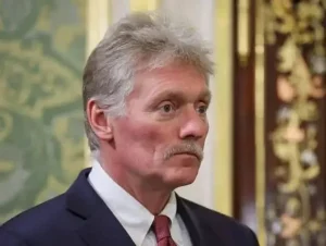 Дмитро Пєсков у темному костюмі з краваткою на тлі орнаментованої стіни