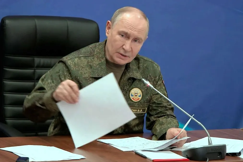 Путін у камуфляжній формі сидить за столом і тримає папери біля мікрофона