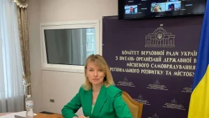Олена Шуляк за столом на тлі банера комітету Ради та прапора України