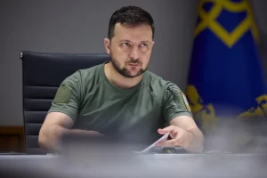 Зеленський у зеленій футболці сидить за столом біля президентського прапора