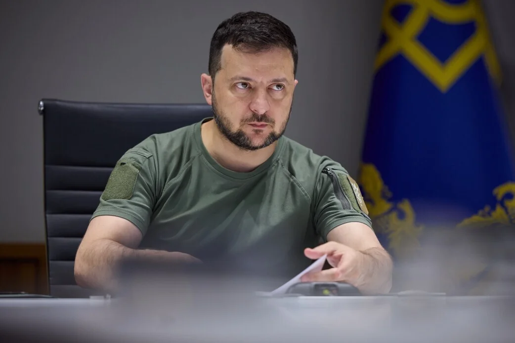Зеленський у зеленій футболці сидить за столом біля президентського прапора